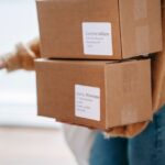 Crop anonymous female wearing casual outfit carrying carton parcels with address labels opening door of post office